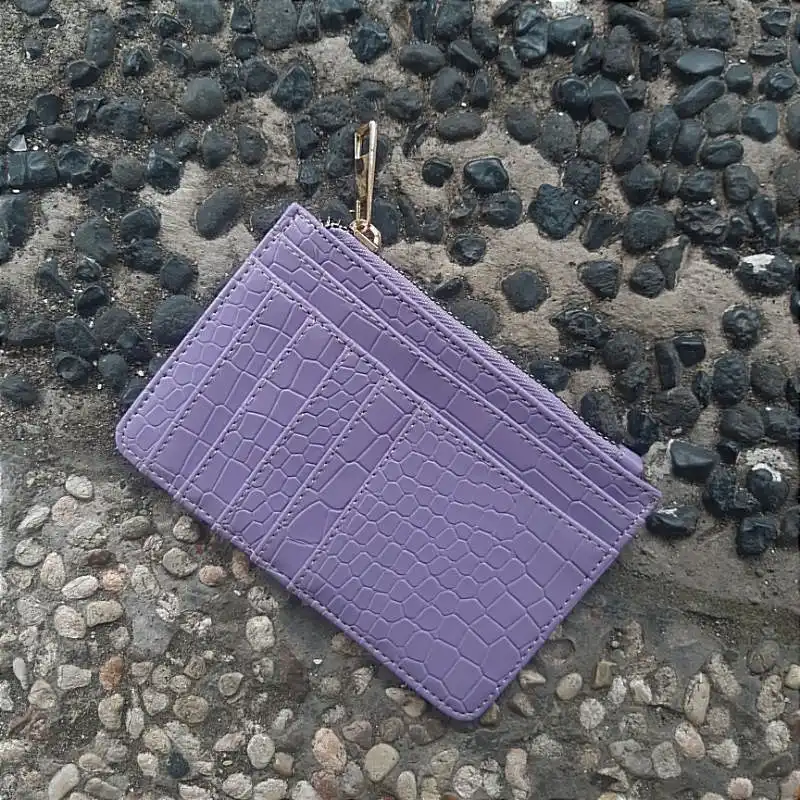 Card holder bertekstur croco warna lilac dengan beberapa slot kartu di sisi depan dan ritsleting di tepi atas dengan penarik logam keemasan, diletakkan di atas lantai batu koral