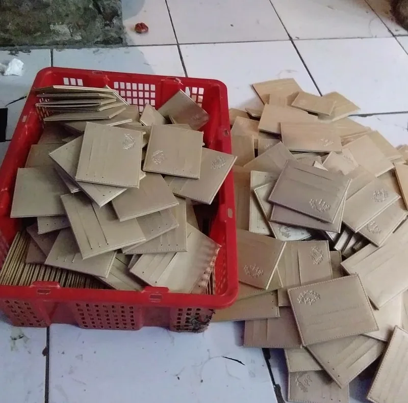 Keranjang plastik merah berisi tumpukan card holder krem yang baru dipotong, dengan card holder lain berserakan di lantai keramik di sebelahnya