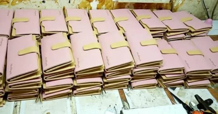 Puluhan dompet lipat berwarna merah muda dengan tali pengunci krem tersusun berderet di atas meja kerja workshop