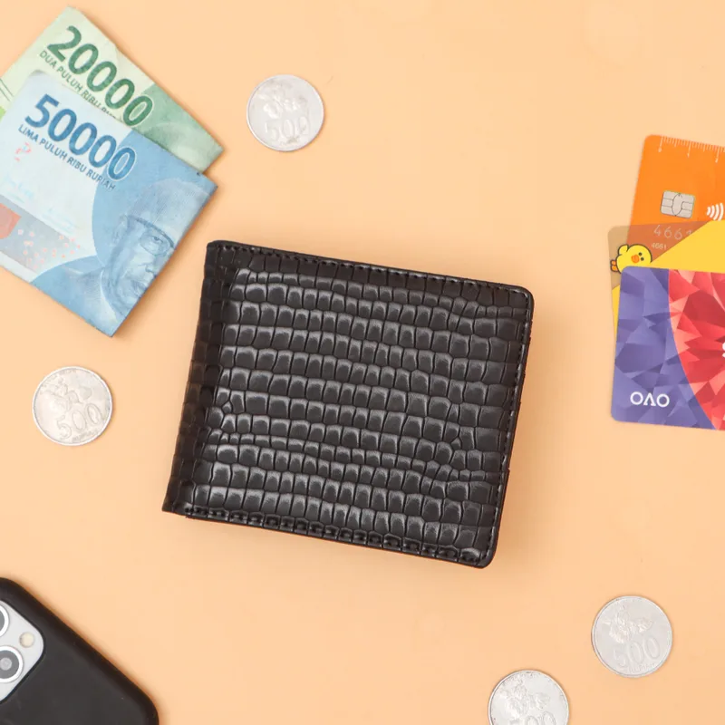 Dompet lipat berwarna hitam dengan tekstur timbul menyerupai sisik, diletakkan di atas latar oranye muda bersama uang kertas, uang logam, dan kartu