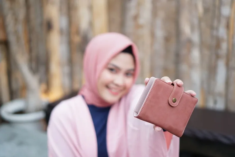 Seorang model berhijab merah muda tersenyum sambil memegang dompet lipat berwarna merah muda dengan tali kancing, dengan latar dinding kayu yang buram