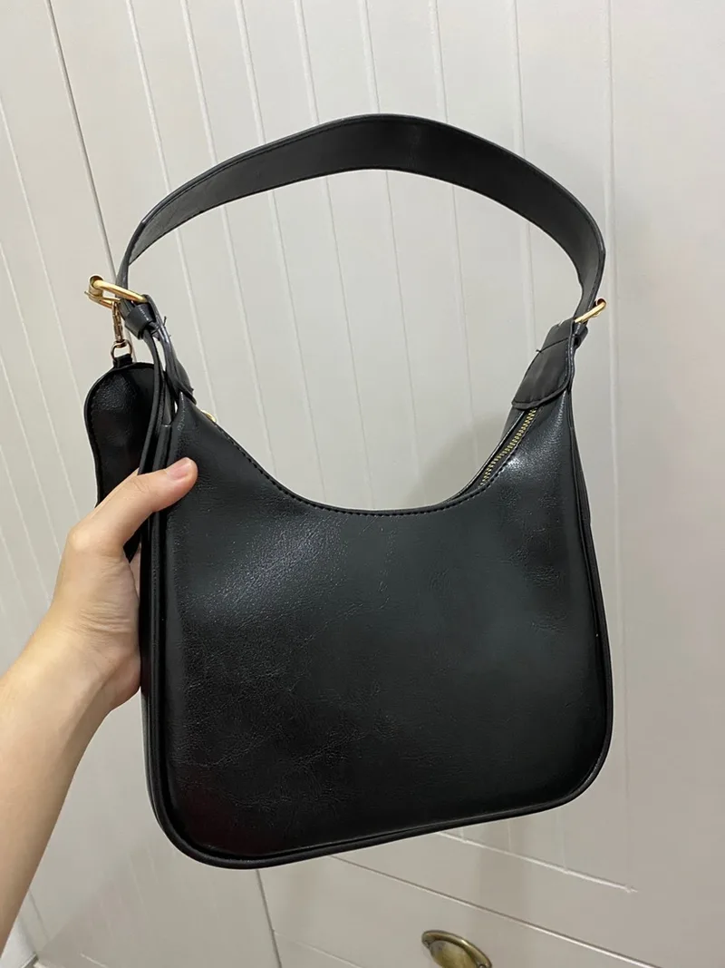 Tas bahu Benita hitam berbahan kulit sintetis dengan tali lebar dan pouch kecil, diangkat dengan satu tangan di depan dinding putih