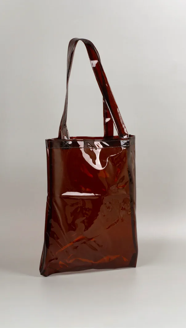 Totebag bening berwarna cokelat kemerahan dengan dua pegangan senada, berdiri di atas alas abu-abu