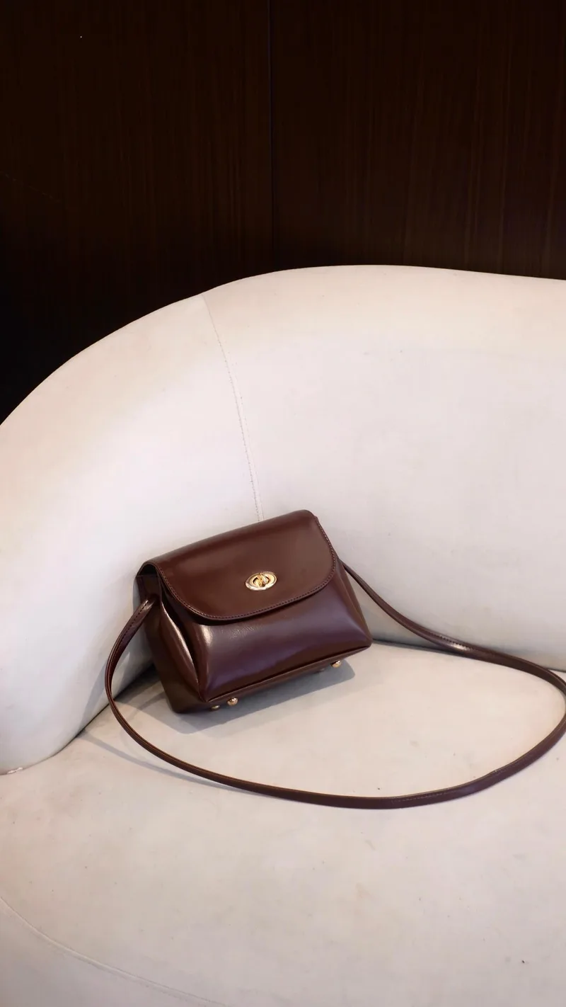 Tas selempang Belly berwarna cokelat tua mengilap dengan tutup depan, kunci putar berwarna emas, dan tali panjang tipis, diletakkan di atas sofa putih