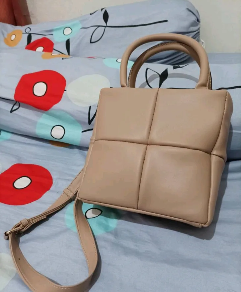 Tas Belva berwarna krem dengan pola kotak empat panel, dua pegangan pendek, dan tali selempang panjang dengan gesper logam, diletakkan di atas sprei bermotif bunga merah