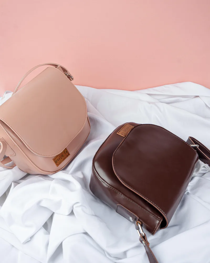Dua tas selempang bertutup setengah lingkaran, satu berwarna merah muda pucat dan satu cokelat tua, dengan label kecil bertuliskan beela co, tergeletak di atas kain putih berlatar dinding merah muda