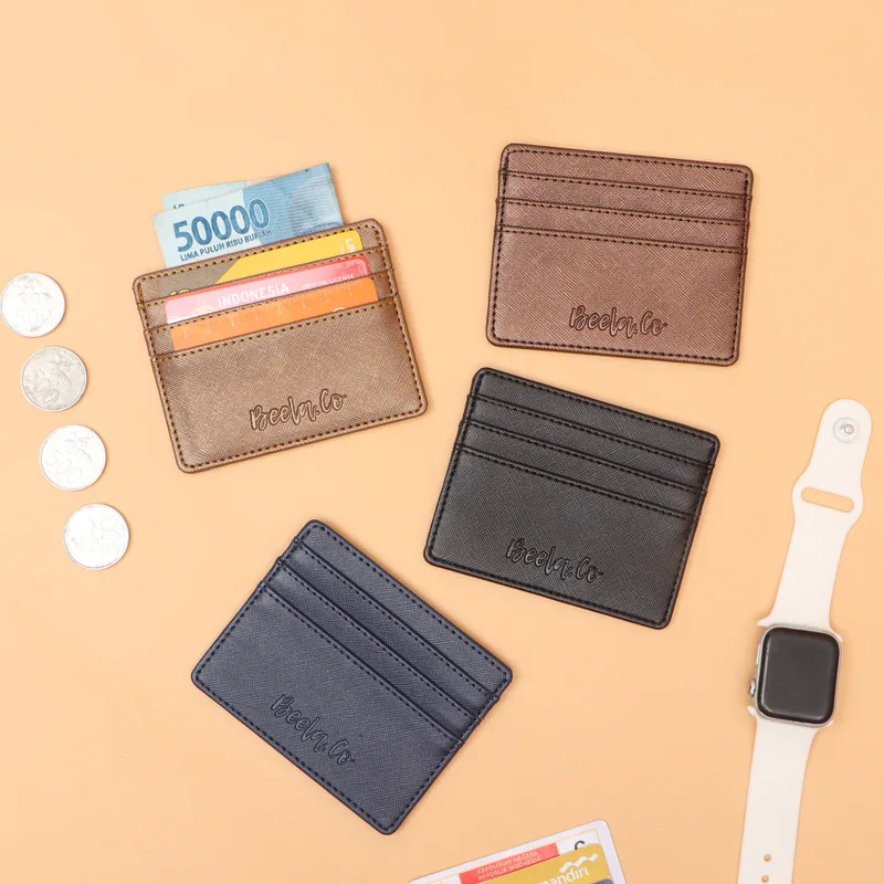 Empat dompet kartu kulit tipis warna cokelat muda, cokelat tua, hitam, dan biru dongker dengan tulisan Beela.Co tercetak, satu berisi kartu dan uang lima puluh ribu rupiah, di atas alas oranye muda bersama koin dan jam tangan