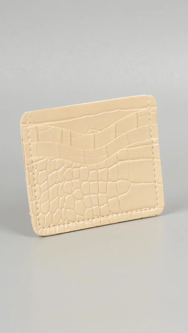 Card holder warna krem dengan tekstur kulit buaya timbul dan slot kartu di bagian depan, berdiri miring di atas latar abu-abu muda