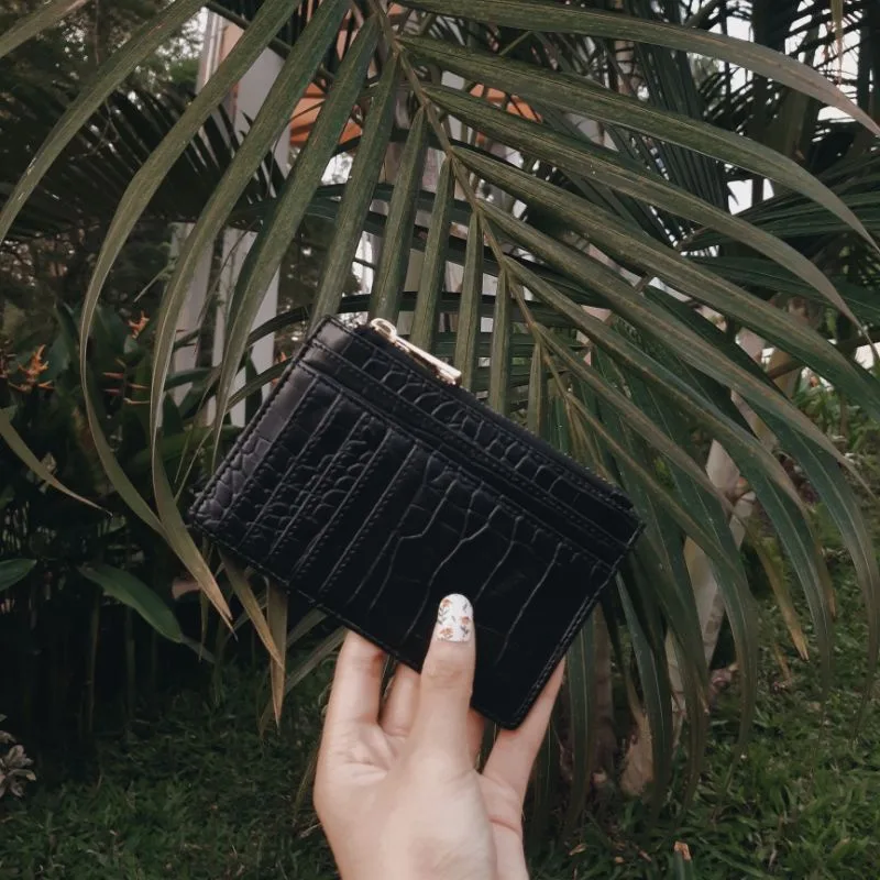 Tangan dengan kuku berhias memegang dompet kartu hitam bertekstur motif croco dengan penarik ritsleting keemasan, berlatar daun palem