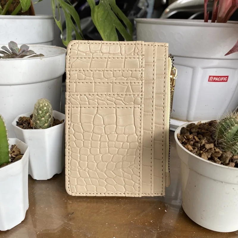 Dompet kartu krem bertekstur motif croco berdiri tegak di atas meja di antara pot kaktus putih, memperlihatkan deretan slot kartu dan ritsleting di sisi kanan