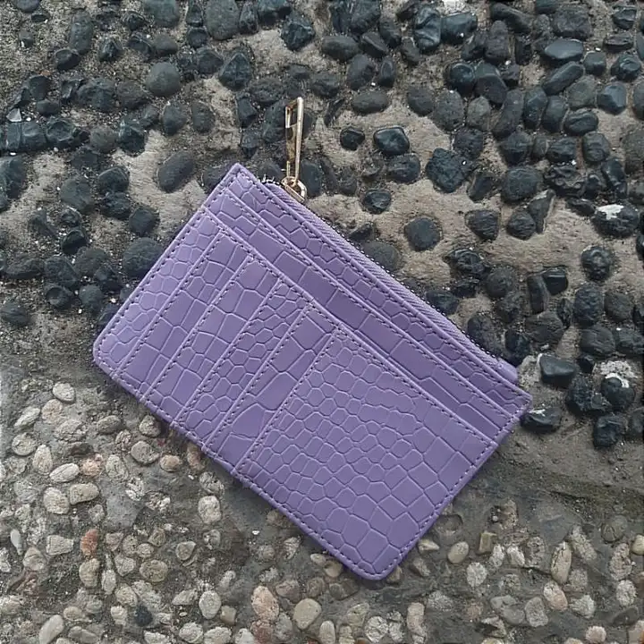 Dompet kartu lilac bertekstur motif croco dengan penarik ritsleting keemasan tergeletak di atas lantai kerikil