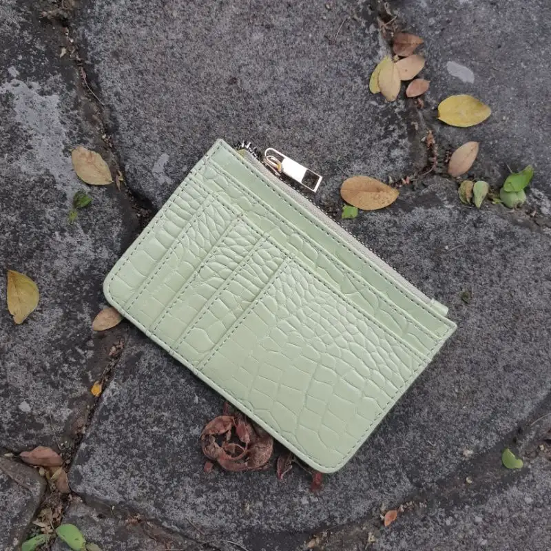 Dompet kartu hijau mint bertekstur motif croco tergeletak di atas batu abu-abu yang dikelilingi daun kering