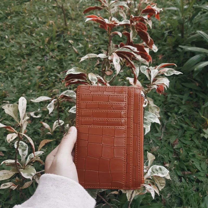 Tangan memegang dompet kartu berwarna oranye bata bertekstur motif croco dengan deretan slot kartu, berlatar tanaman berdaun merah dan rumput