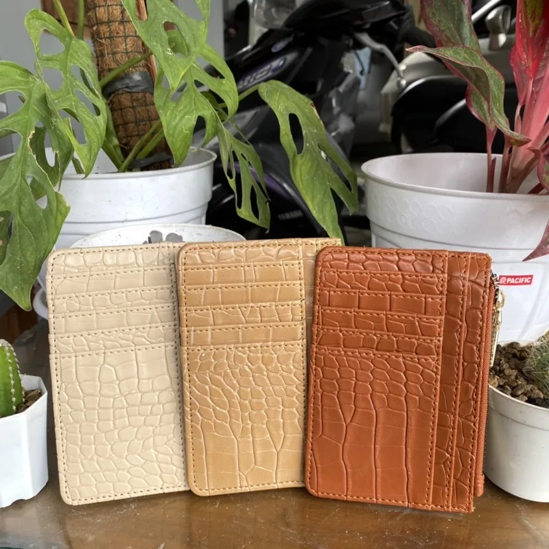 Tiga dompet kartu bertekstur kulit buaya warna krem, cokelat muda, dan cokelat oranye berdiri berjajar di atas meja dengan latar pot tanaman