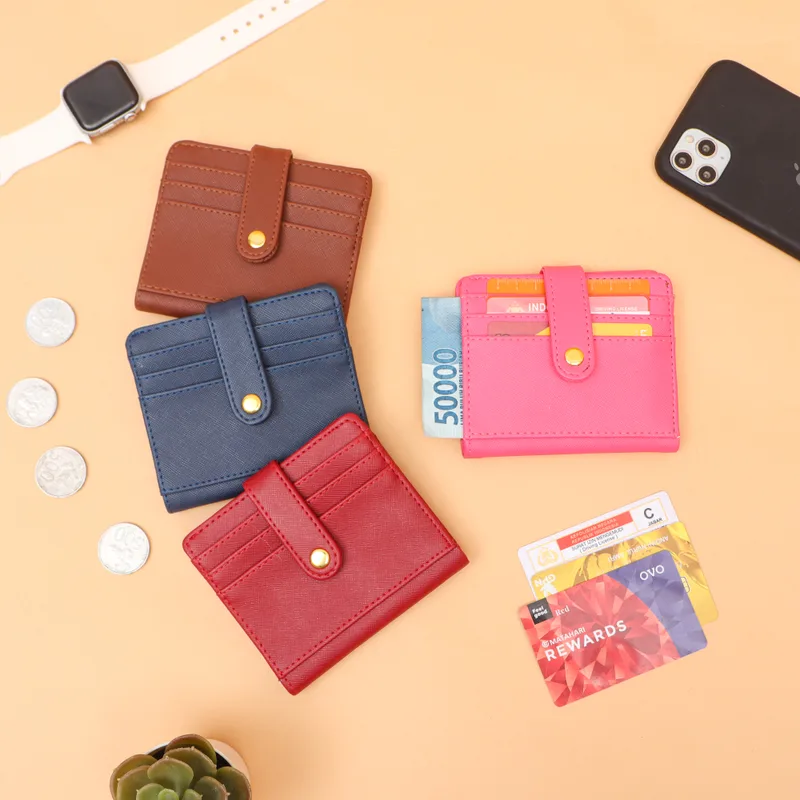 Empat dompet kartu kecil berkancing emas warna cokelat, biru dongker, merah, dan merah muda di atas alas oranye muda, bersama koin, kartu, uang lima puluh ribu rupiah, jam tangan, dan ponsel