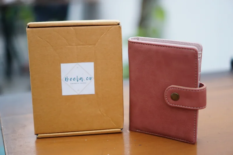 Dompet lipat berwarna merah bata dengan tali berkancing logam berdiri di samping kotak kemasan kardus cokelat berlabel Beela.co di atas meja kayu