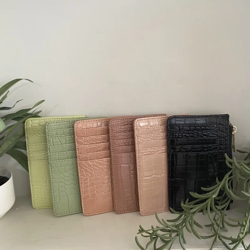 Enam dompet kartu bertekstur croco berdiri berjajar dengan warna hijau muda, hijau sage, peach, cokelat muda, nude, dan hitam di depan dinding abu-abu