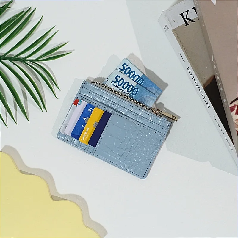 Dompet kartu bertekstur croco warna biru muda berisi beberapa kartu di slot depan dan dua lembar uang lima puluh ribu di kantong ritsleting