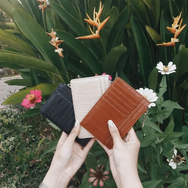 Dua tangan memegang tiga dompet kartu bertekstur croco warna hitam, krem, dan cokelat dengan latar tanaman dan bunga