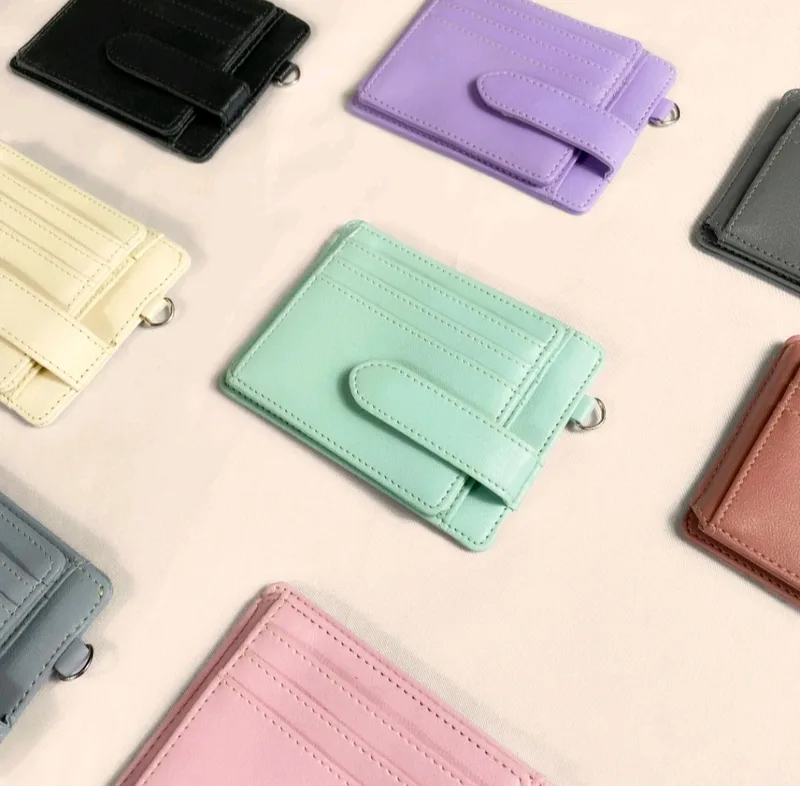 Beberapa card holder warna ungu, hijau mint, krem, abu-abu, dan hitam dengan tali pengunci dan ring logam kecil, tersebar di atas latar merah muda