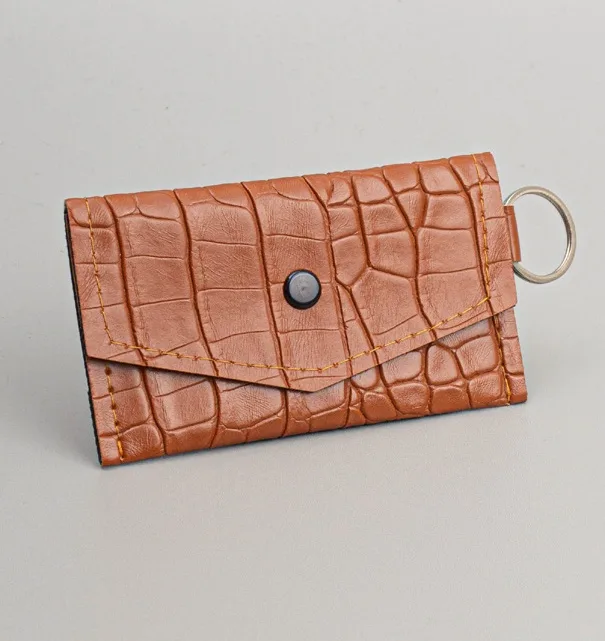 Gantungan kunci model dompet STNK cokelat bertekstur motif croco dengan tutup berkancing hitam dan ring logam di sisi kanan, di atas latar abu-abu