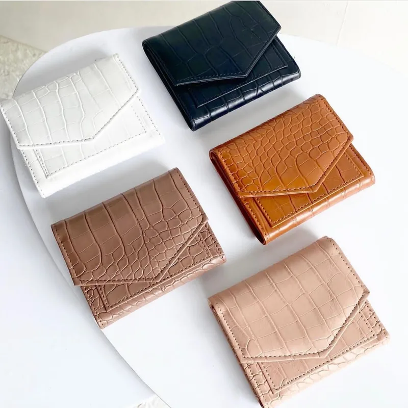 Lima dompet lipat kecil bertekstur motif kulit buaya berwarna putih, hitam, cokelat tan, cokelat susu, dan krem dengan penutup lipat bersudut, tersusun di atas meja putih