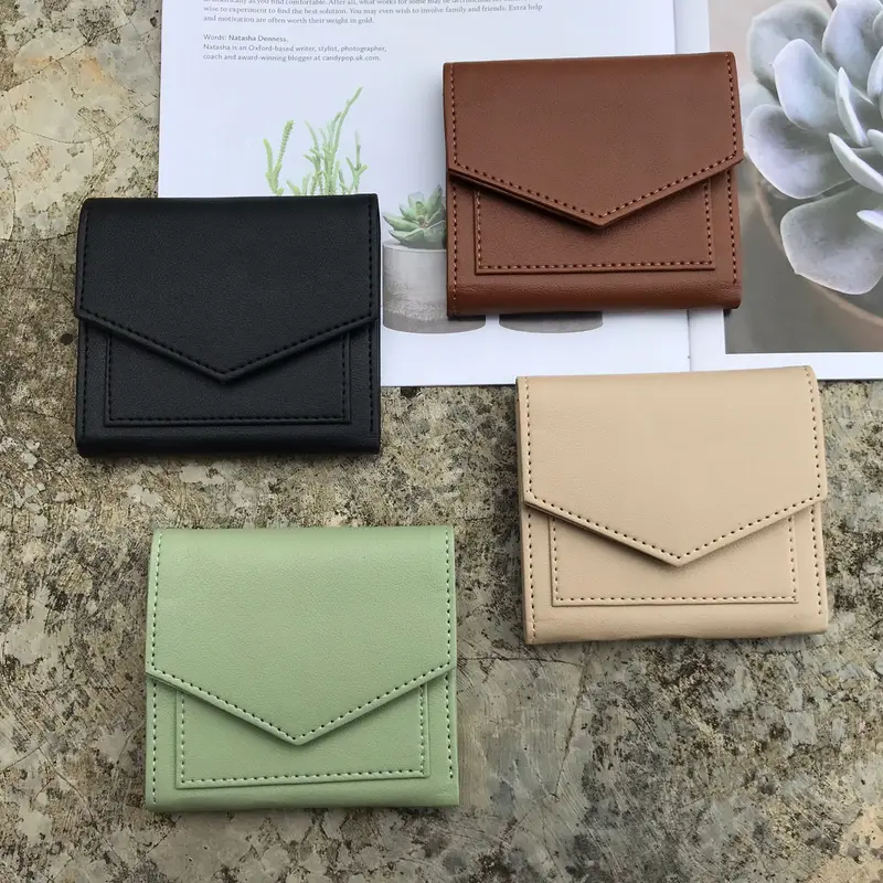 Empat dompet lipat kecil polos berpenutup bersudut warna hitam, cokelat, krem, dan hijau sage tersusun di atas permukaan batu bercorak, dengan selembar kertas bergambar tanaman dan tanaman sukulen di latar