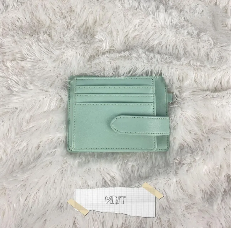 Card holder berwarna hijau mint dengan beberapa slot kartu dan tali penutup di sisi kanan, diletakkan di atas karpet bulu putih dengan label kertas bertuliskan MINT