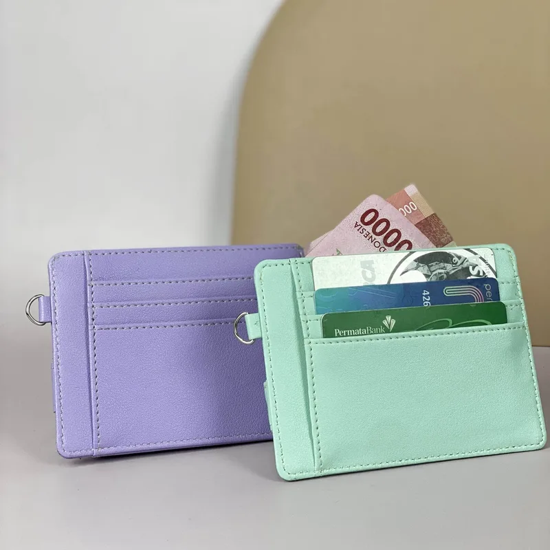 Dua card holder berdiri di atas meja putih, satu berwarna ungu lavender dengan cincin logam di sisi dan satu berwarna hijau mint berisi beberapa kartu dan lembaran uang
