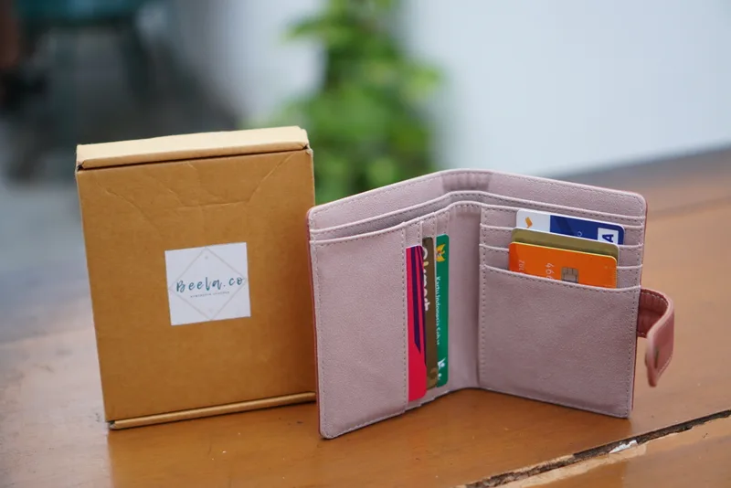 Dompet lipat warna merah muda keunguan terbuka berisi kartu, berdiri di samping kotak kemasan cokelat berlabel, di atas meja kayu