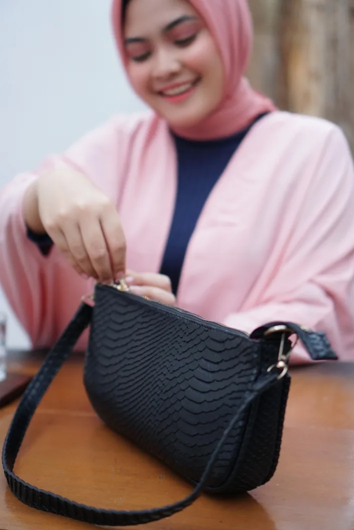 Perempuan berhijab merah muda tersenyum sambil membuka tas bahu hitam bertekstur sisik di atas meja kayu