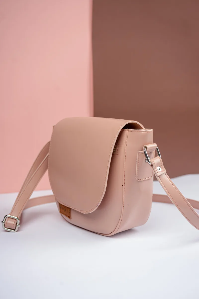 Tas selempang merah muda pucat bertutup setengah lingkaran dengan tali panjang dan label kecil di bagian depan, di atas latar merah muda dan cokelat