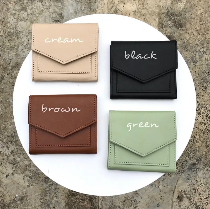 Empat dompet lipat bertutup amplop berwarna krem, hitam, cokelat, dan hijau dengan tulisan nama warna, diletakkan di atas piring putih bundar di permukaan batu