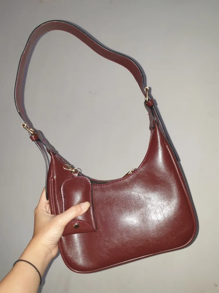 Tangan memegang tas bahu cokelat kemerahan berbentuk setengah bulan dengan tali bahu panjang dan dompet kecil yang menggantung di samping tas