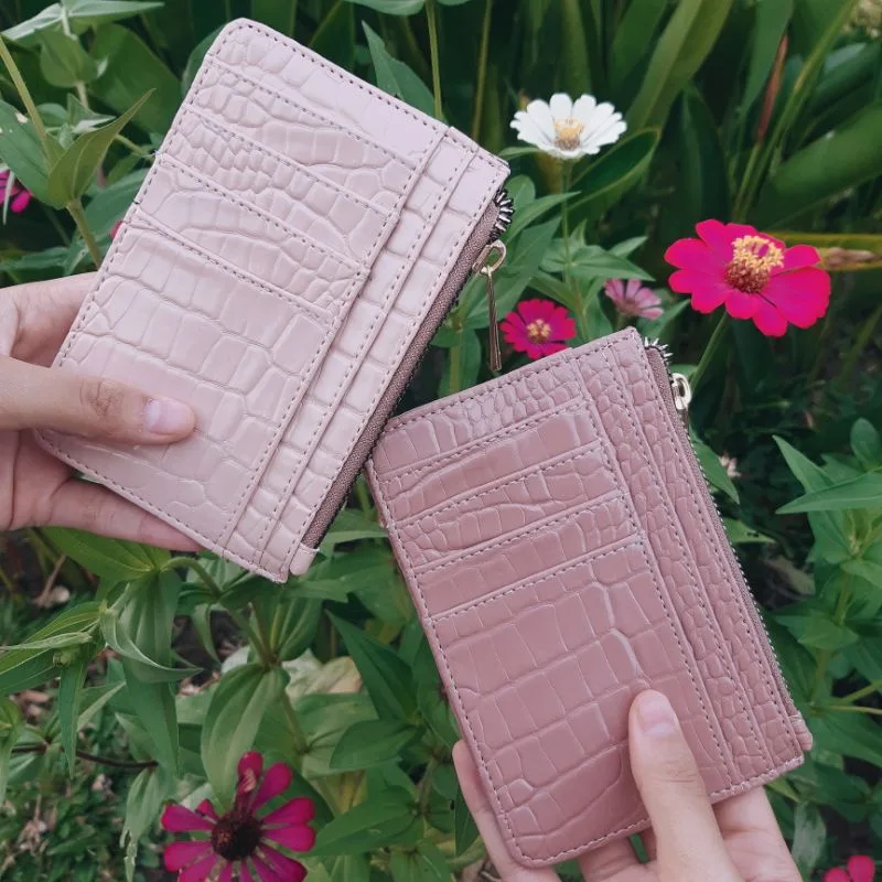 Dua tangan memegang dompet kartu bertekstur croco warna merah muda pucat dan merah muda dengan ritsleting di bagian atas, berlatar bunga zinnia dan daun hijau