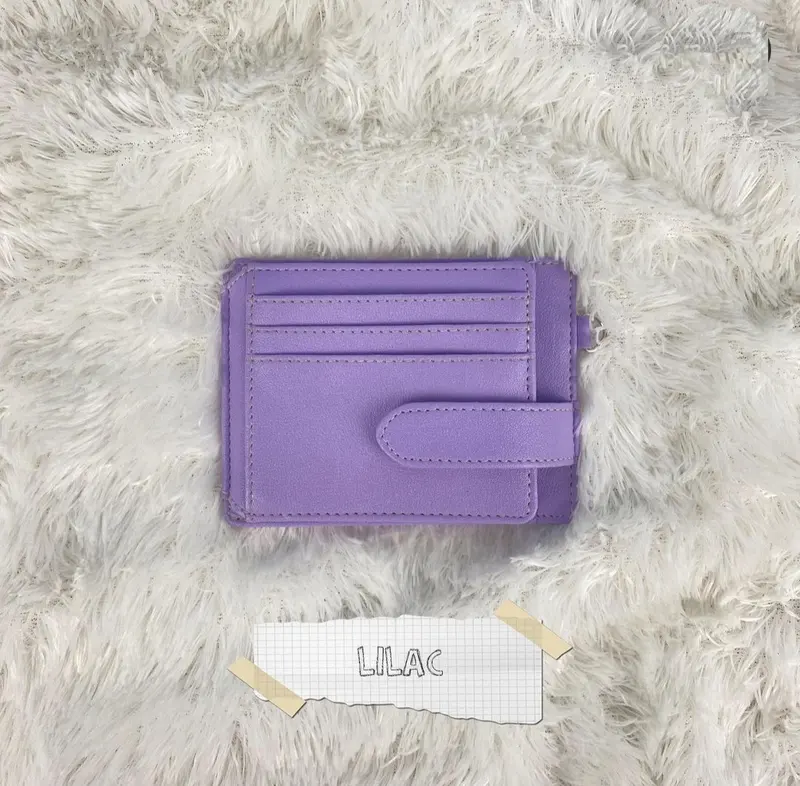 Card holder kecil warna lilac atau ungu muda dengan slot kartu bertingkat dan tali pengunci samping di atas alas bulu putih, dengan label kertas bertuliskan lilac