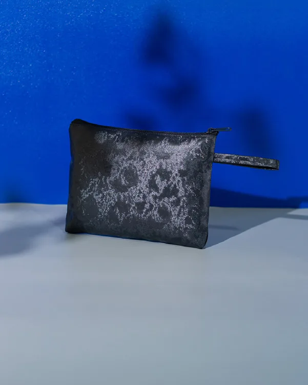 Pouch beritsleting hitam dengan corak abu-abu kebiruan seperti marmer dan tali genggam di sisi kanan, berdiri di depan latar biru dengan bayangan daun