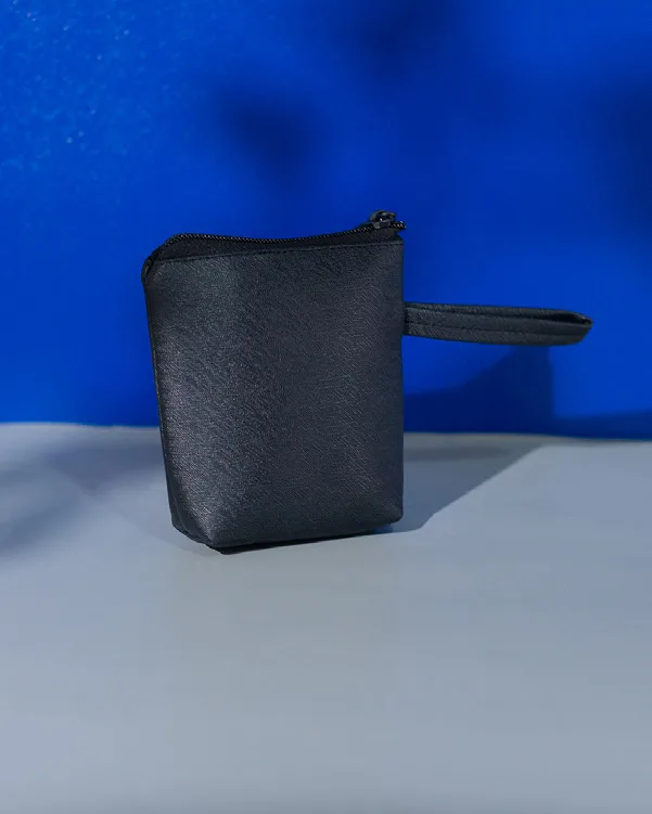 Pouch kecil berwarna hitam dengan tekstur halus berkilau, ritsleting di bagian atas, dan tali genggam di sisi kanan, berdiri di depan latar biru