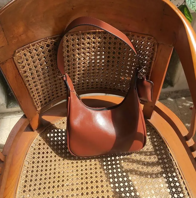 Tas bahu berwarna cokelat kemerahan dengan tali lebar dan pouch kecil yang tergantung, diletakkan di atas kursi kayu beranyaman rotan yang terkena cahaya matahari