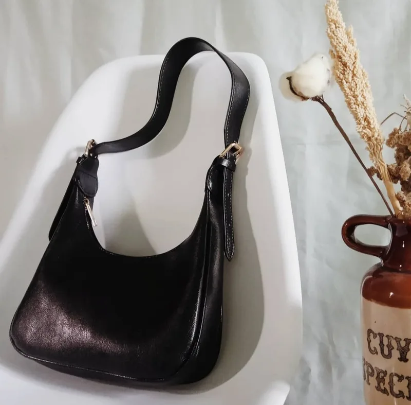 Tas bahu hitam mengilap dengan tali lebar dan pengait logam keemasan, diletakkan di kursi putih dengan hiasan bunga kapas kering di sampingnya