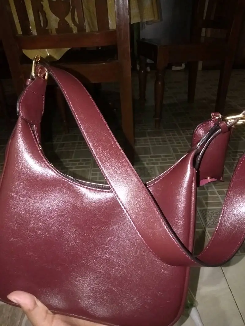 Tas bahu berwarna merah marun dengan permukaan halus mengilap dan tali lebar, dipegang dekat kamera di dalam ruangan dengan kursi kayu di latar belakang