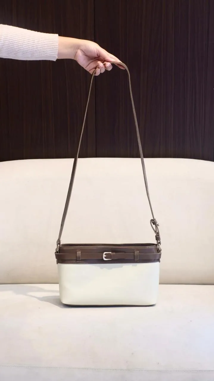 Tas bahu berbadan putih gading dengan bagian atas dan tali berwarna cokelat tua serta gesper kecil di depan, dijinjing tangan di atas sofa putih dengan latar gelap