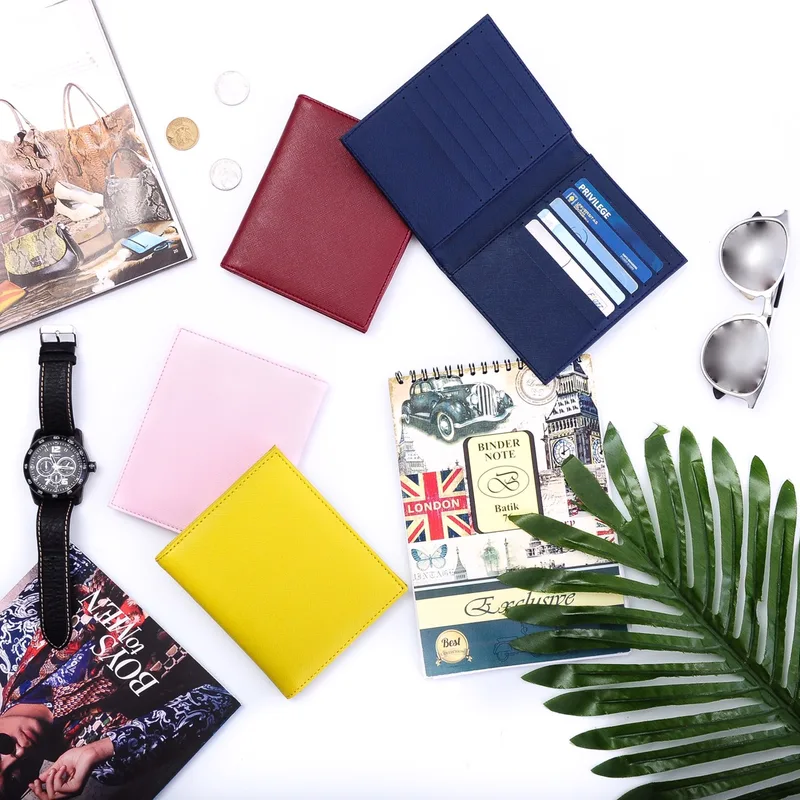 Dompet lipat warna marun, merah muda pucat, dan kuning tertutup, serta satu dompet biru dongker terbuka memperlihatkan deretan slot berisi kartu, di atas meja putih bersama jam tangan, buku catatan, kacamata hitam, dan daun palem