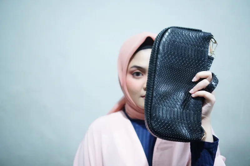 Perempuan berhijab merah muda memegang tas hitam bertekstur sisik di depan wajahnya sehingga motif sisik dan pengait logam di ujung tas terlihat dari dekat
