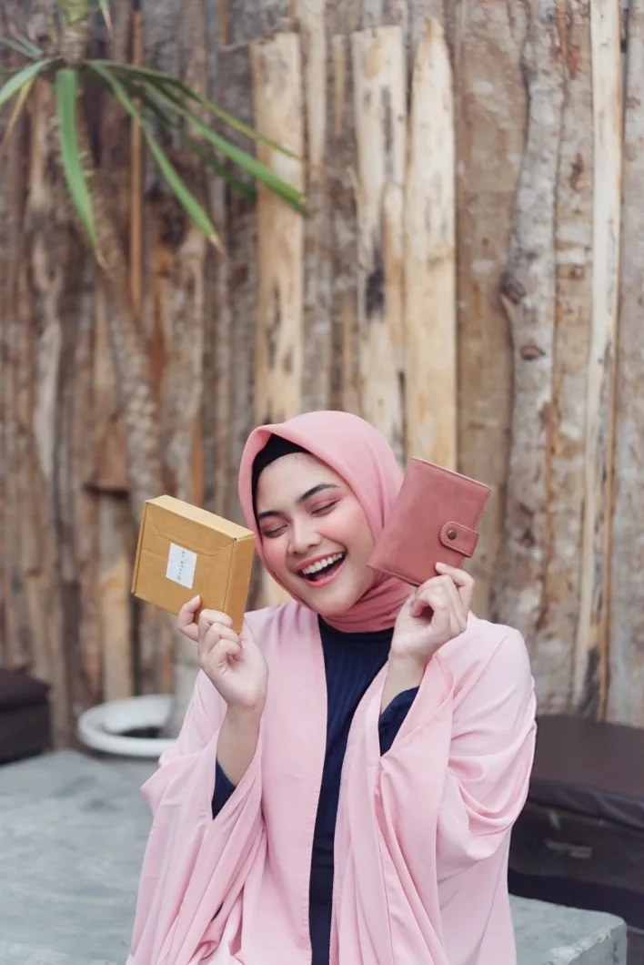 Perempuan berhijab merah muda tertawa sambil memegang kotak kemasan kardus cokelat di tangan kanan dan dompet merah muda berkancing jepret di tangan kiri, dengan latar dinding batang kayu