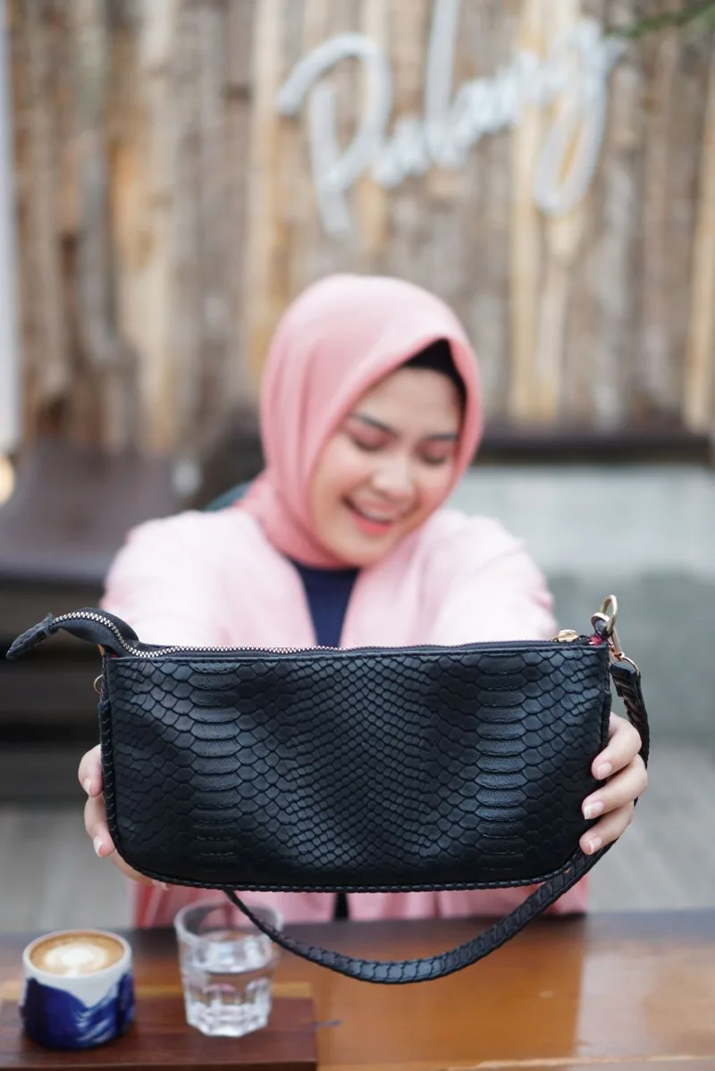 Perempuan berhijab merah muda tersenyum sambil menyodorkan tas bahu hitam bertekstur sisik dengan ritsleting dan pengait logam ke arah kamera, di depan meja kayu dengan secangkir kopi dan gelas air