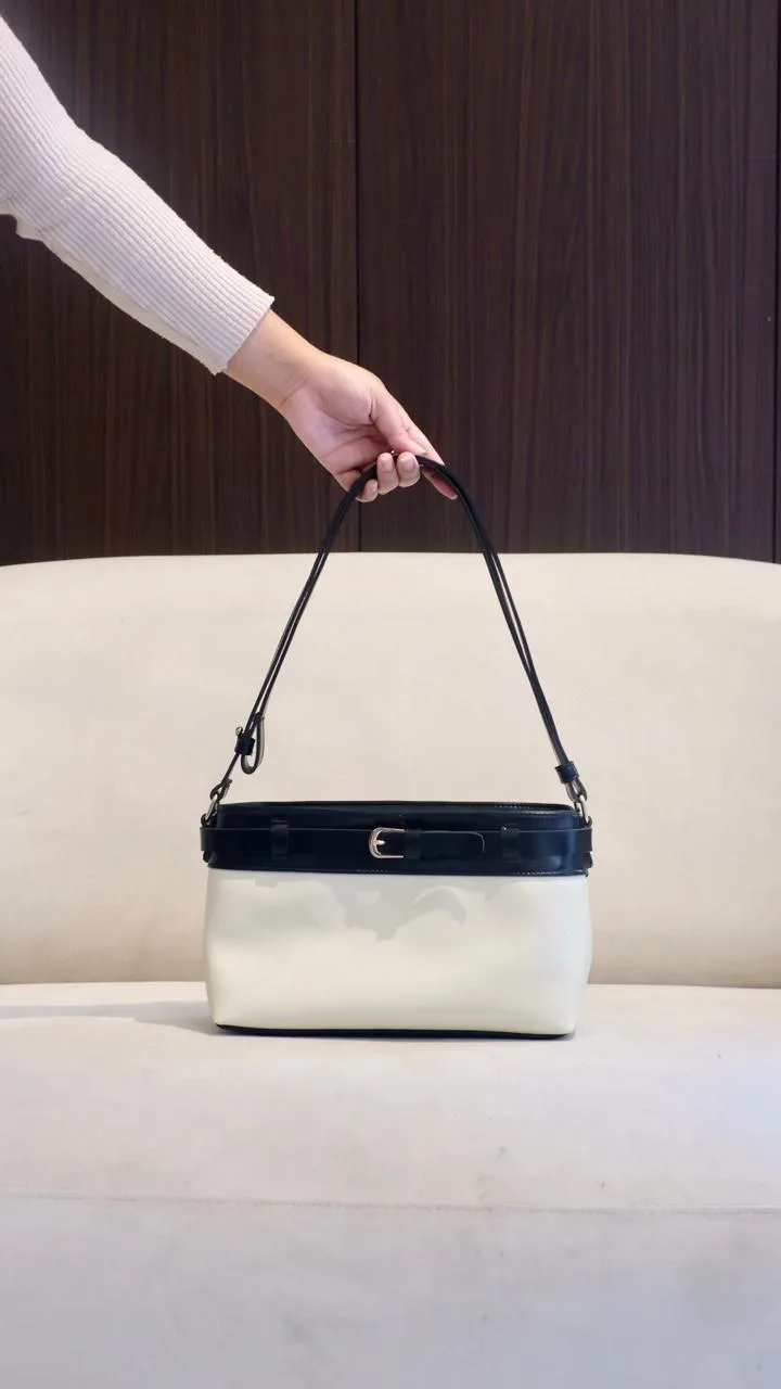 Tangan menjinjing tas bahu dua warna dengan badan putih gading dan bagian atas hitam bergesper kecil, tali hitam terhubung ke badan tas lewat pengait logam, di atas sofa krem