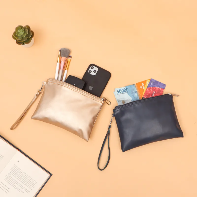 Dua pouch bertali tangan, satu warna emas metalik berisi kuas rias dan satu biru dongker berisi kartu, di atas latar oranye muda bersama ponsel dan buku