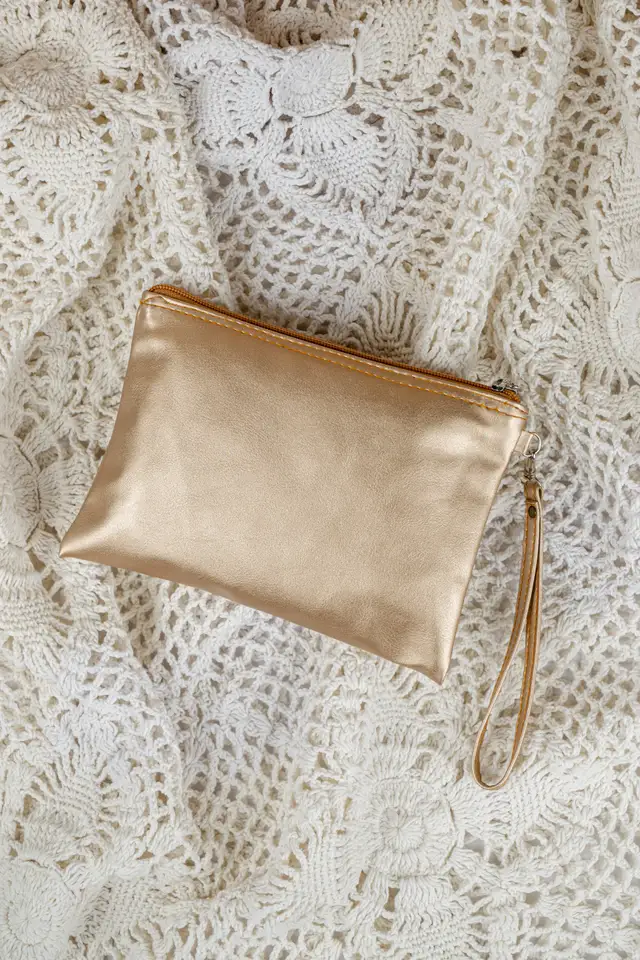 Pouch pipih warna emas metalik dengan tali tangan tipis diletakkan di atas kain renda putih