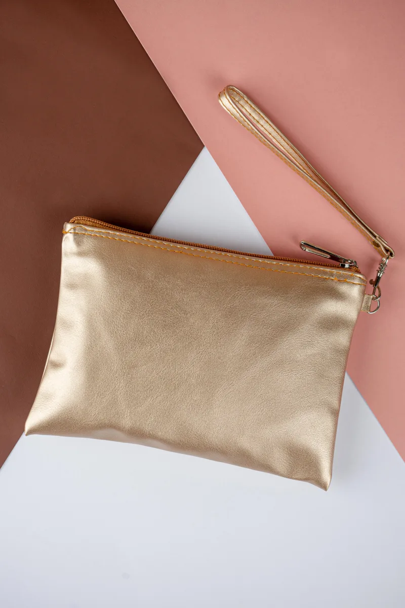 Pouch pipih warna emas metalik dengan tali tangan tipis di atas latar merah muda dan cokelat